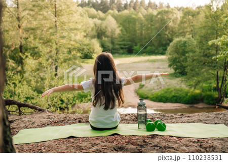 Brunette little girl with arms wide open sitting on green rug on sandy ground. Dumbbells and bottle lying near athlete. 110238531