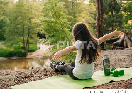 Female kid with arms wide open sitting on mat on sandy hill in front of river with bottle of water and dumbbells near. 110238532