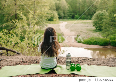 Little female sportsman sit on mat on hill in front of lake and enjoy nature. Dumbbells and bottle lying near child. 110238533