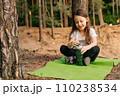 Delightful smiling female kid sitting on green sporty rug outdoor and opening bottle of water that holding in hands. 110238534
