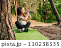 Little girl with dark long hair sitting on green rug in nature and drinking water from bottle, resting after workout. 110238535