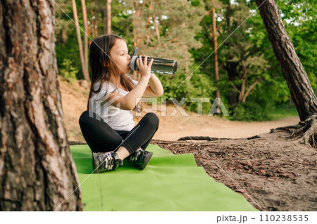 Little girl with dark long hair sitting on green rug in nature and drinking water from bottle, resting after workout. 110238535