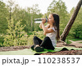 Little girl with dark long hair sitting on green rug in nature and drinking water from bottle, resting after workout. 110238537