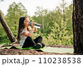 Little girl with dark long hair sitting on green rug in nature and drinking water from bottle, resting after workout. 110238538