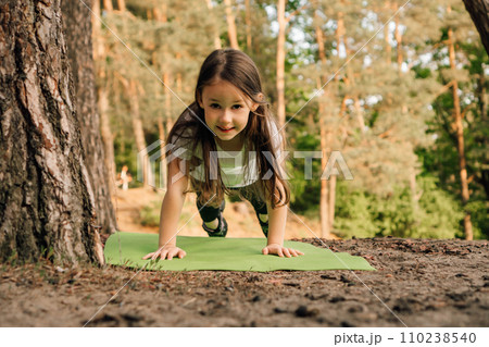 Loveable girl with flowing hair doing plank exercise on green mat outdoor. Junior female athlete working out in nature. 110238540
