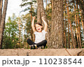 Delightful girl sitting on ground among trees with closed eyes and raised arms. Happy child enjoying activity outdoor. 110238544