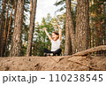 Happy female kid with open mouth sitting on ground in forest with raised arms. Sporty girl enjoying hiking in nature. 110238545