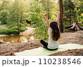 Sporty girl with long hair sitting on green mat, rising arms while enjoying nature. Child resting in front of river. 110238546