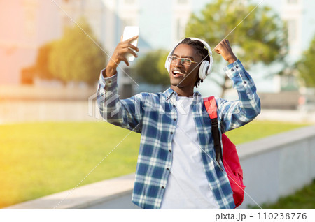 Joyful African guy wearing headphones holding smartphone video calling outside Joyful African guy wearing headphones holding smartphone video calling outside 110238776