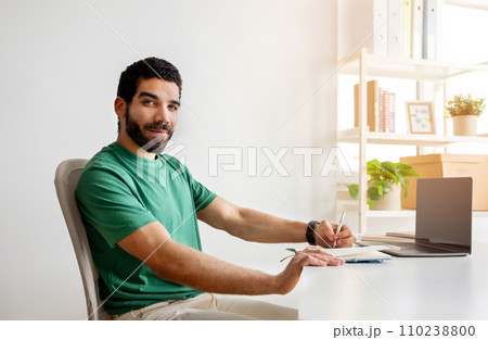 Smiling bearded man with glasses in a green shirt sitting at a desk 110238800
