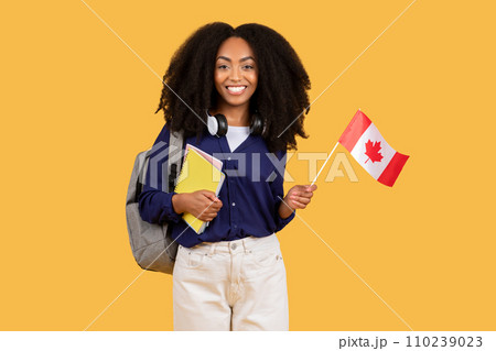 Happy black student with backpack, copybooks, holding Canada flag on yellow background, symbolizing language education 110239023