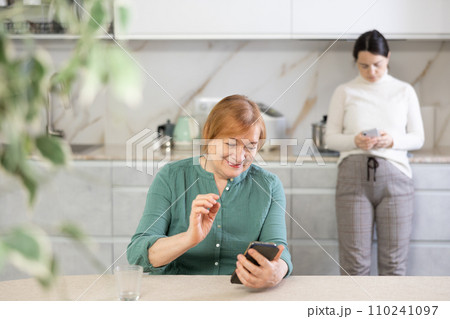 Positive mature woman and girl in kitchen with phone in hand communicates in social network Positive mature woman and girl in kitchen with phone in hand communicates in social network 110241097