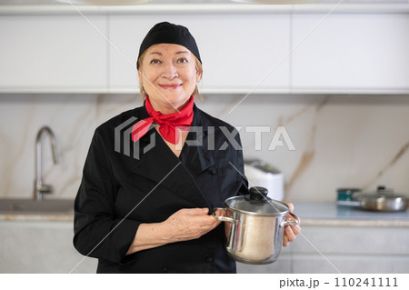 Elderly woman cook in chef hat and black uniform stands in kitchen and holds stainless pan Elderly woman cook in chef hat and black uniform stands in kitchen and holds stainless pan 110241111
