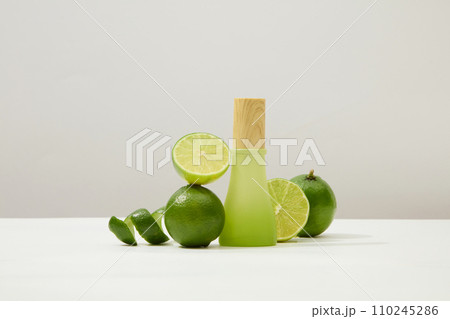 Front view of a green glass bottle with wooden lid, next to fresh lemon on a minimalist white background. Vitamin C in lemon helps restore damaged skin very quickly. 110245286