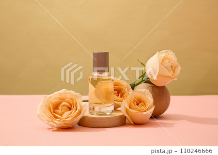 A labelless transparent perfume bottle is placed on a wooden platform with fresh roses. Natural cosmetics concept for advertising. Pink surface with brown background. 110246666