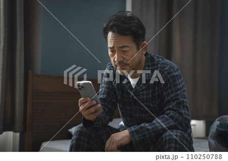 ベッドに座ってスマートフォンの画面を見つめる中年男性 ベッドに座ってスマートフォンの画面を見つめる中年男性 110250788