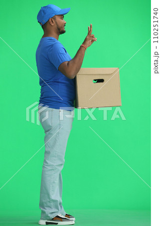 A male deliveryman with a box, on a green background, in full height, shows a victory sign, profile A male deliveryman with a box, on a green background, in full height, shows a victory sign, profile 110251740