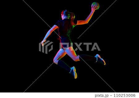 Fit young woman engaged in intense handball training, perfecting her throwing and catching abilities against black background in neon light, filter. 110253006