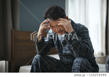 ベッドに座って頭を抱える50代半ばの男性 110254349