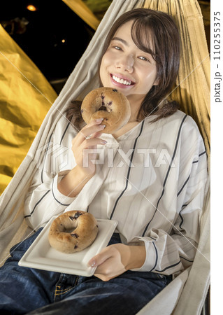 室内キャンプ、ハンモックでベーグルを食べる若い女性 110255375