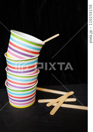A stack of colorful striped paper cups and wooden stirrers on a black background. The cups are adorned with horizontal stripes in various bright colors including red, green blue and yellow. 110258156