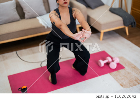 Overweight woman enjoying a fitness workout at home. Fat, plump woman and squatting on an exercise mat in the living room Overweight woman enjoying a fitness workout at home. Fat, plump woman and squatting on an exercise mat in the living room 110262042