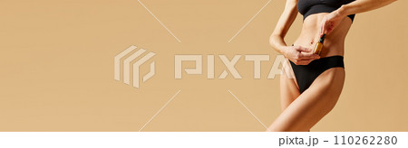 Cropped image of female body, relief belly in black cotton underwear against beige background. Model holding moisturizing cream Cropped image of female body, relief belly in black cotton underwear against beige background. Model holding moisturizing cream 110262280