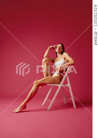 Full-length image of young woman with slim fit healthy body sitting on chair in white cotton underwear against pink studio background Full-length image of young woman with slim fit healthy body sitting on chair in white cotton underwear against pink studio background 110262329