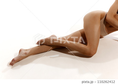 Cropped image of slender, tanned, smooth female legs. Model posing in beige cotton underwear against white studio background. Depilation, epilation 110262412