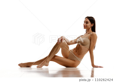 Beautiful young woman with relief slim fit body sitting on floor, posing in beige cotton underwear against white studio background Beautiful young woman with relief slim fit body sitting on floor, posing in beige cotton underwear against white studio background 110262424
