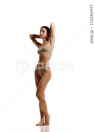 Full-length image of young woman with slim relief tanned body standing in beige cotton underwear against white studio background Full-length image of young woman with slim relief tanned body standing in beige cotton underwear against white studio background 110262447