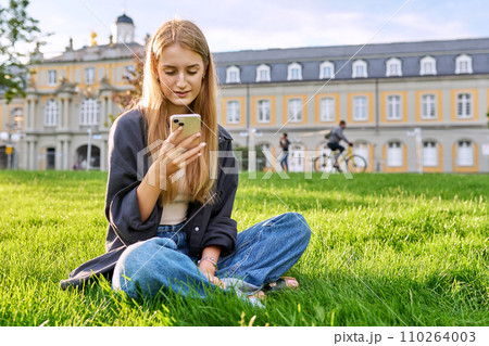 Young smiling female using smartphone sitting on grass, lawn in sunset light Young smiling female using smartphone sitting on grass, lawn in sunset light 110264003