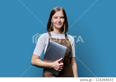 Portrait of young woman in apron holding laptop on blue background 110264033