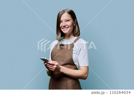 Portrait of young woman in apron holding smartphone in hands on blue background 110264034