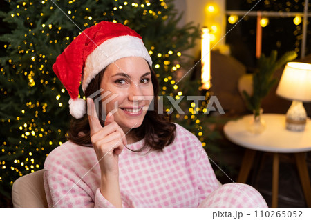 An attractive young woman in cozy pajamas is sitting at home on a chair in a red Christmas hat, smiling and holding her index finger up. An attractive young woman in cozy pajamas is sitting at home on a chair in a red Christmas hat, smiling and holding her index finger up. 110265052