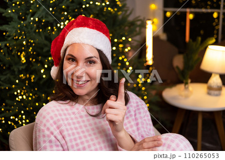 An attractive young woman in cozy pajamas is sitting at home on a chair in a red Christmas hat, smiling and holding her index finger up. An attractive young woman in cozy pajamas is sitting at home on a chair in a red Christmas hat, smiling and holding her index finger up. 110265053