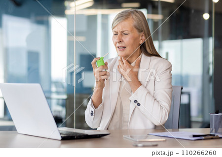 Senior business woman sitting in the office at the desk and holding her throat, feeling severe pain, using medical aerosol. Senior business woman sitting in the office at the desk and holding her throat, feeling severe pain, using medical aerosol. 110266260