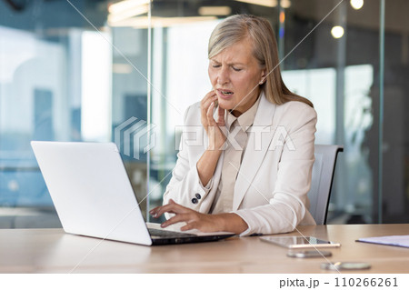 An older gray-haired businesswoman is sitting at a desk in the office, working on a laptop, holding her hand to her cheek, suffering from a severe toothache. An older gray-haired businesswoman is sitting at a desk in the office, working on a laptop, holding her hand to her cheek, suffering from a severe toothache. 110266261