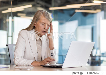 Senior gray-haired businesswoman working in the office, sitting at the desk, holding her head with her hand and suffering from a headache. Senior gray-haired businesswoman working in the office, sitting at the desk, holding her head with her hand and suffering from a headache. 110266262