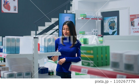 Smiling asian customer amidst pharmacy shelves, looking at medicine packages, trying to find suitable remedy for troubling ailments. Joyful cilent holding shopping bag, browsing through products 110267776