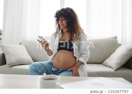 Pregnant woman blogger sits on the couch at home and takes pictures of herself on the phone, selfie and video call, consultation with the doctor online, pregnancy management, freelance work Pregnant woman blogger sits on the couch at home and takes pictures of herself on the phone, selfie and video call, consultation with the doctor online, pregnancy management, freelance work 110267787