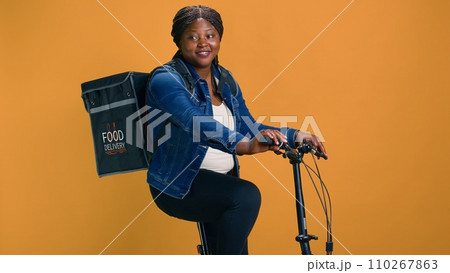 Young and active courier using modern bicycle to deliver package with precision to the city office. African american lady with backpack working as professional delivery person. 110267863