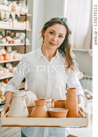 A woman master of a pottery workshop carries clay products for firing. Potter with an assortment of ceramic dishes in a bright workshop A woman master of a pottery workshop carries clay products for firing. Potter with an assortment of ceramic dishes in a bright workshop 110270111