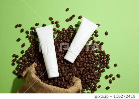 On a green background, coffee beans falling from a burlap bag and two empty cosmetic tubes are decorated. Top view, mockup scene for advertising. Natural ingredient for skin care and body care On a green background, coffee beans falling from a burlap bag and two empty cosmetic tubes are decorated. Top view, mockup scene for advertising. Natural ingredient for skin care and body care 110272419