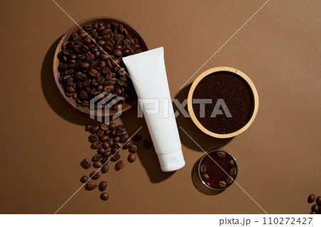 Minimal scene for advertising cosmetic with natural ingredient. Top view of a white plastic bottle without label decorated with wooden bowl of coffee beans and grounds on brown background Minimal scene for advertising cosmetic with natural ingredient. Top view of a white plastic bottle without label decorated with wooden bowl of coffee beans and grounds on brown background 110272427