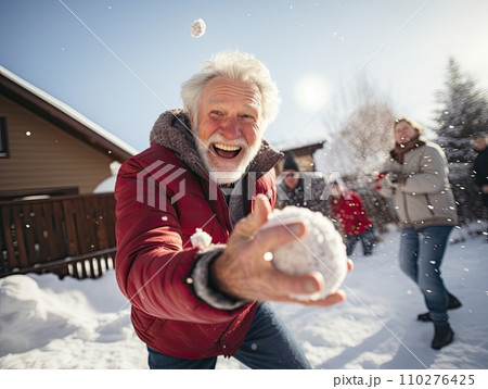 Joyful elderly man in a red jacket takes a selfie while playfully throwing a snowball. Another man in a Santa hat is in the background in a snowy setting Joyful elderly man in a red jacket takes a selfie while playfully throwing a snowball. Another man in a Santa hat is in the background in a snowy setting 110276425