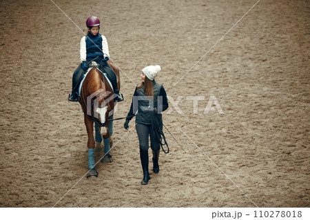 Aerial, top view. Female teacher, instructor teaching little girl, kid horseback riding. Careful ride, outdoor lesson Aerial, top view. Female teacher, instructor teaching little girl, kid horseback riding. Careful ride, outdoor lesson 110278018