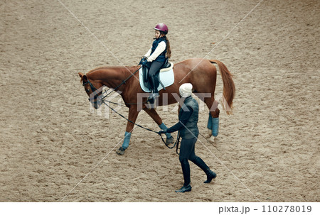 Aerial, top view. Female teacher, instructor teaching little girl, kid horseback riding. Careful ride, outdoor lesson Aerial, top view. Female teacher, instructor teaching little girl, kid horseback riding. Careful ride, outdoor lesson 110278019