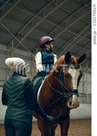 Female instructor teaching little girl, child horseback rising. Kid practicing trail riding, dressage Female instructor teaching little girl, child horseback rising. Kid practicing trail riding, dressage 110278029