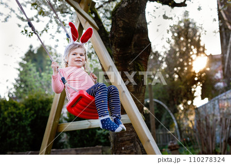 Happy beautiful little toddler girl with red Easter bunny ears having fun on swing in domestic garden. Cute healthy child swinging under blooming trees on sunny spring day. Baby laughing and crying 110278324